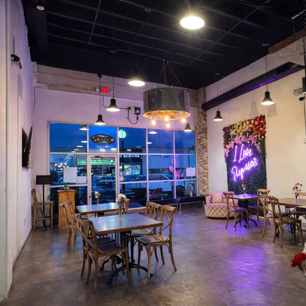 Restaurant interior with tables, chairs, a large window, and a neon "I Love Pupusas" sign on a floral wall.
