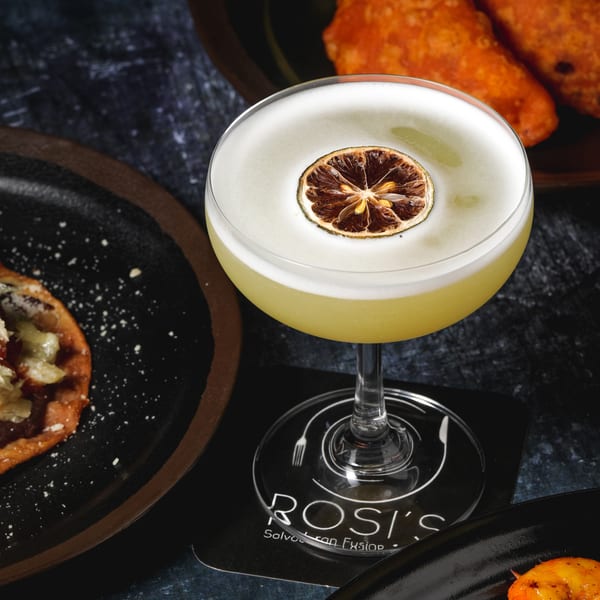 Yellow cocktail with a dried lime garnish on a Rosi's Salvadoran Fusion coaster, with food nearby.