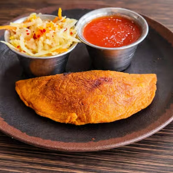 A golden-brown empanada with two metal cups of coleslaw and red sauce on a dark plate.