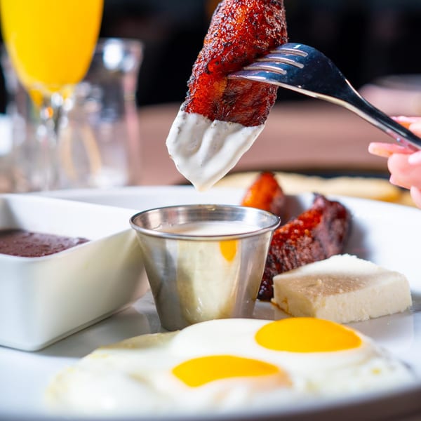 Hand dipping a crispy plantain into creamy white sauce, with fried eggs and cheese on a plate.