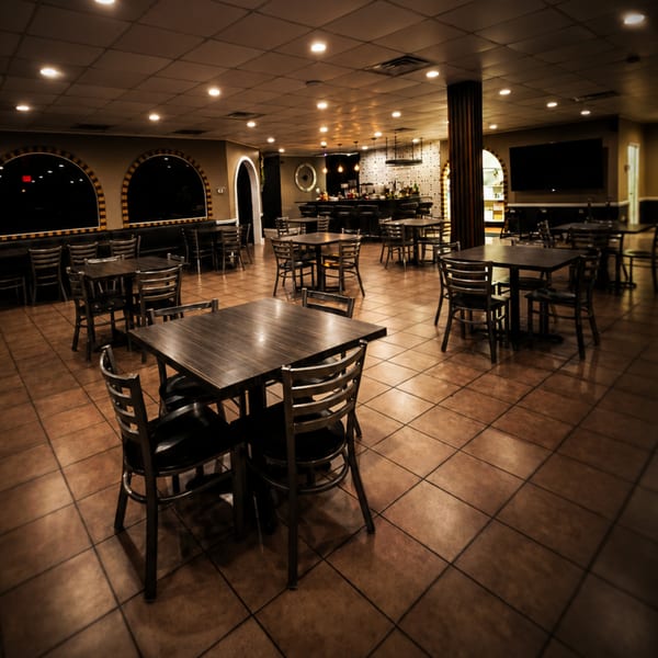 Warmly lit, empty restaurant dining room with dark tables, chairs, and a bar.