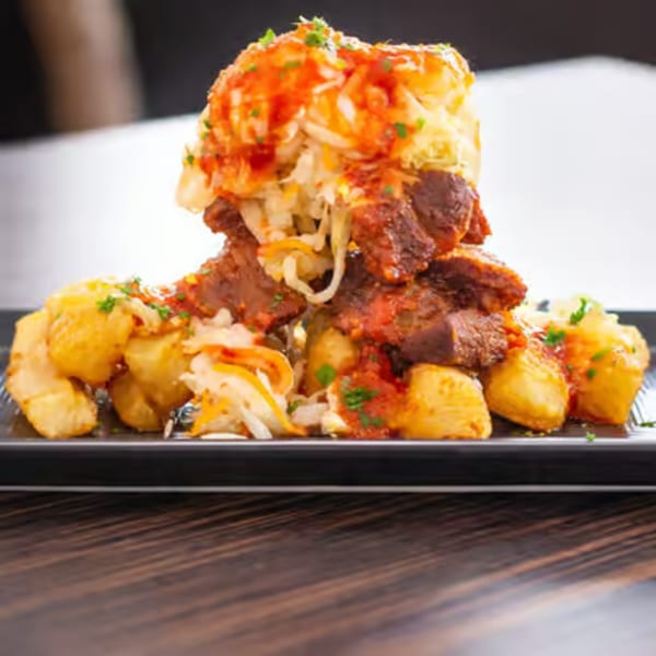 A towering dish of meat, potatoes, and coleslaw, drizzled with red sauce and garnished with herbs.
