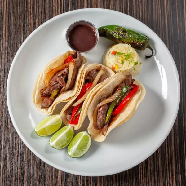 Three steak fajita tacos with peppers and onions, served with rice, lime wedges, and sauce.