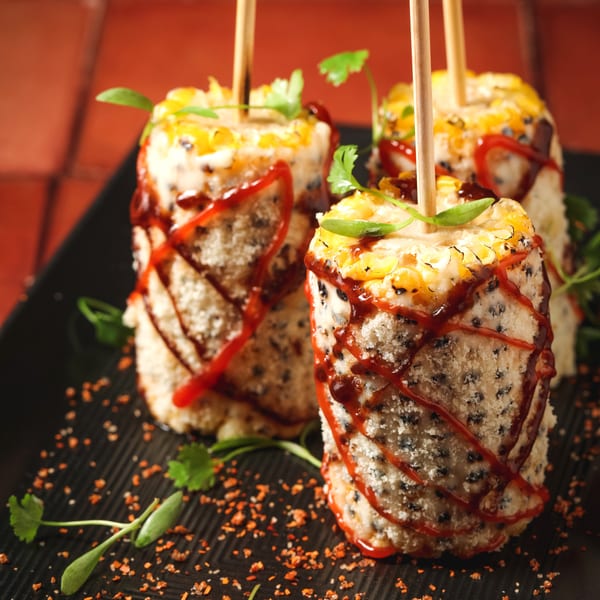 Three skewered corn on the cob pieces, coated in white cheese, black specks, red sauce, and fresh herbs.