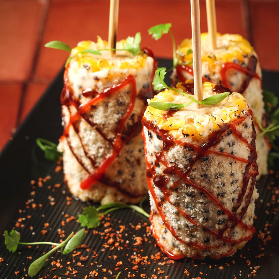 Three skewered corn on the cob pieces, coated in white cheese, black specks, red sauce, and fresh herbs.