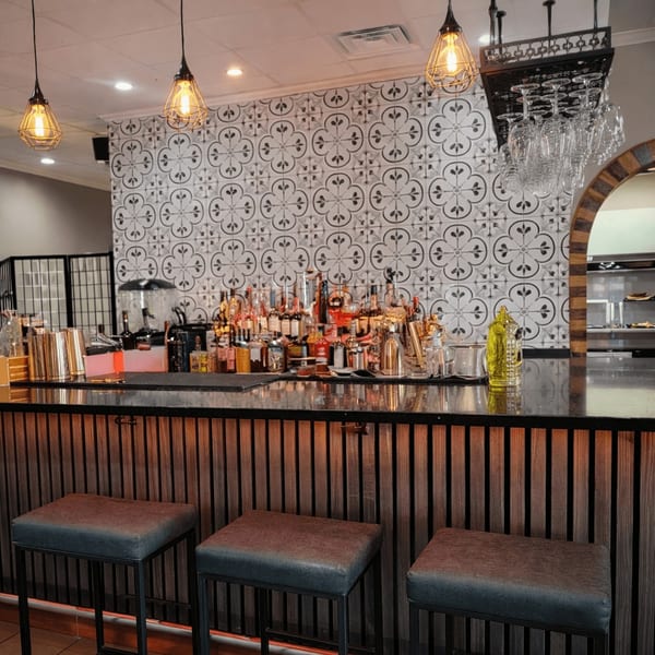 A well-stocked bar with bottles, shakers, and three bar stools, against a patterned tile wall.