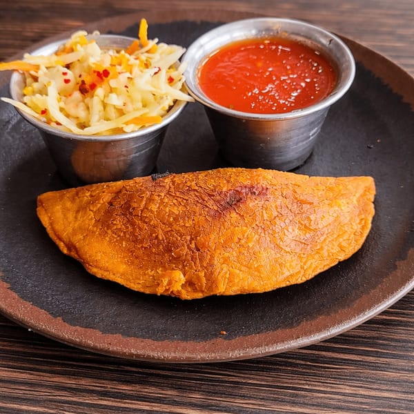A golden-brown empanada with two metal cups of coleslaw and red sauce on a dark plate.