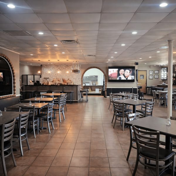 Spacious restaurant dining room with many empty tables, a bar area, and a large TV screen.